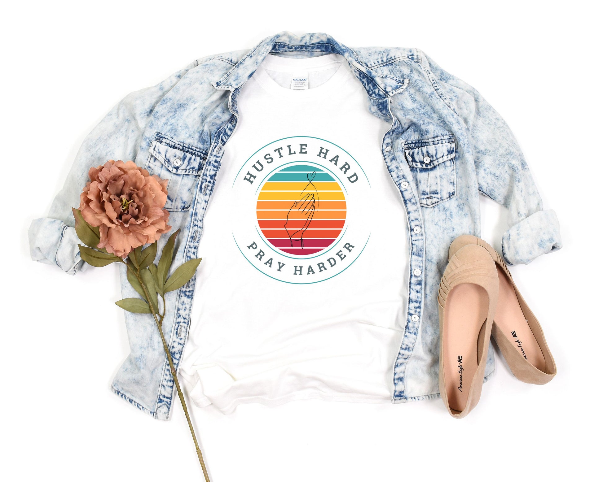 White crew neck t-shirt with the text 'HUSTLE HARD PRAY HARDER' printed on the front, surrounded by a multicolored sunset circle, placed on a wooden surface with a jacket, shoes, and foliage beside it.