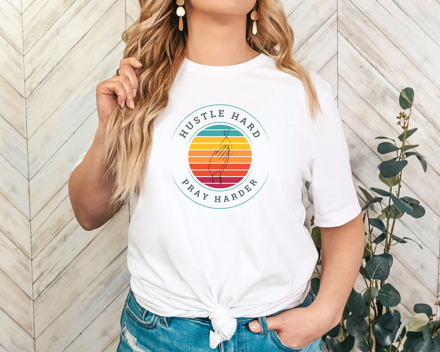 White crew neck t-shirt with the text 'HUSTLE HARD PRAY HARDER' printed on the front, surrounded by a multicolored sunset circle, worn by a model.