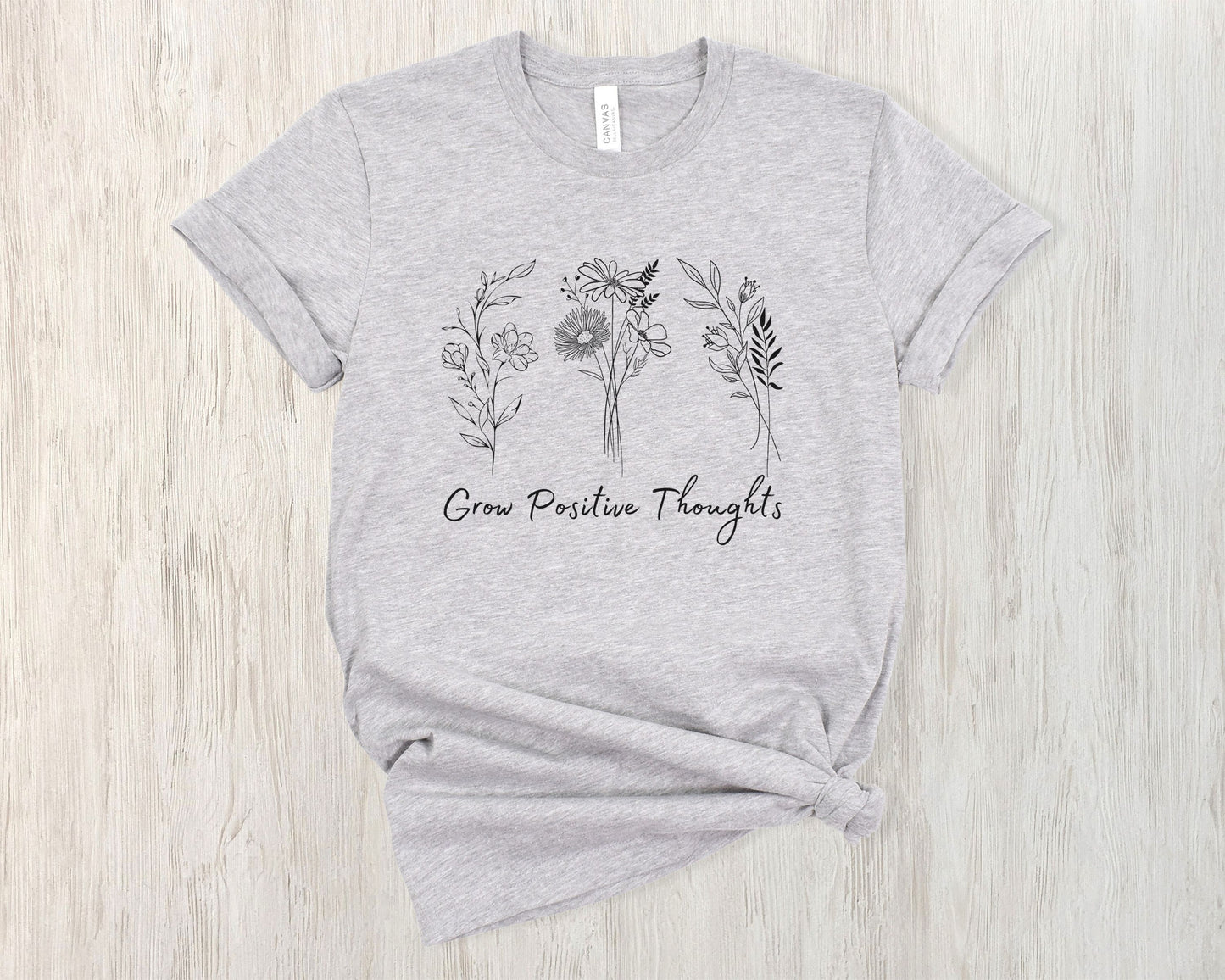 "Grow Positive Thoughts" t-shirt featuring black botanical floral illustrations and an uplifting message in elegant script. Available in white, ash, peach, red, and pink. Soft, lightweight fabric, perfect for spreading positivity and encouragement.