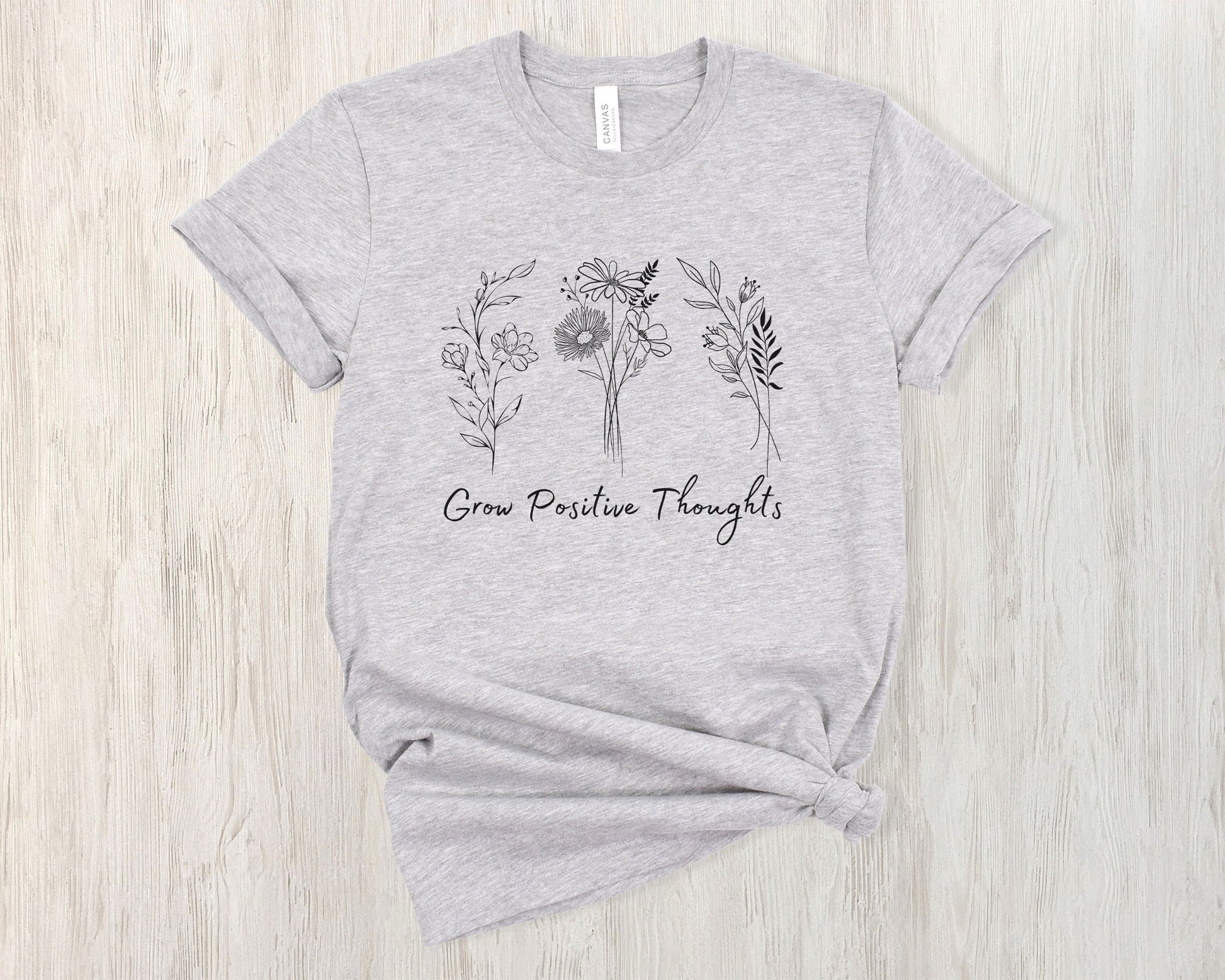 "Grow Positive Thoughts" t-shirt featuring black botanical floral illustrations and an uplifting message in elegant script. Available in white, ash, peach, red, and pink. Soft, lightweight fabric, perfect for spreading positivity and encouragement.