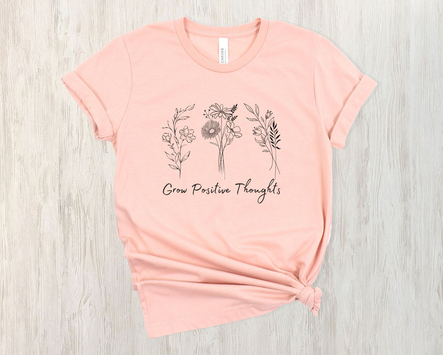 "Grow Positive Thoughts" t-shirt featuring black botanical floral illustrations and an uplifting message in elegant script. Available in white, ash, peach, red, and pink. Soft, lightweight fabric, perfect for spreading positivity and encouragement.