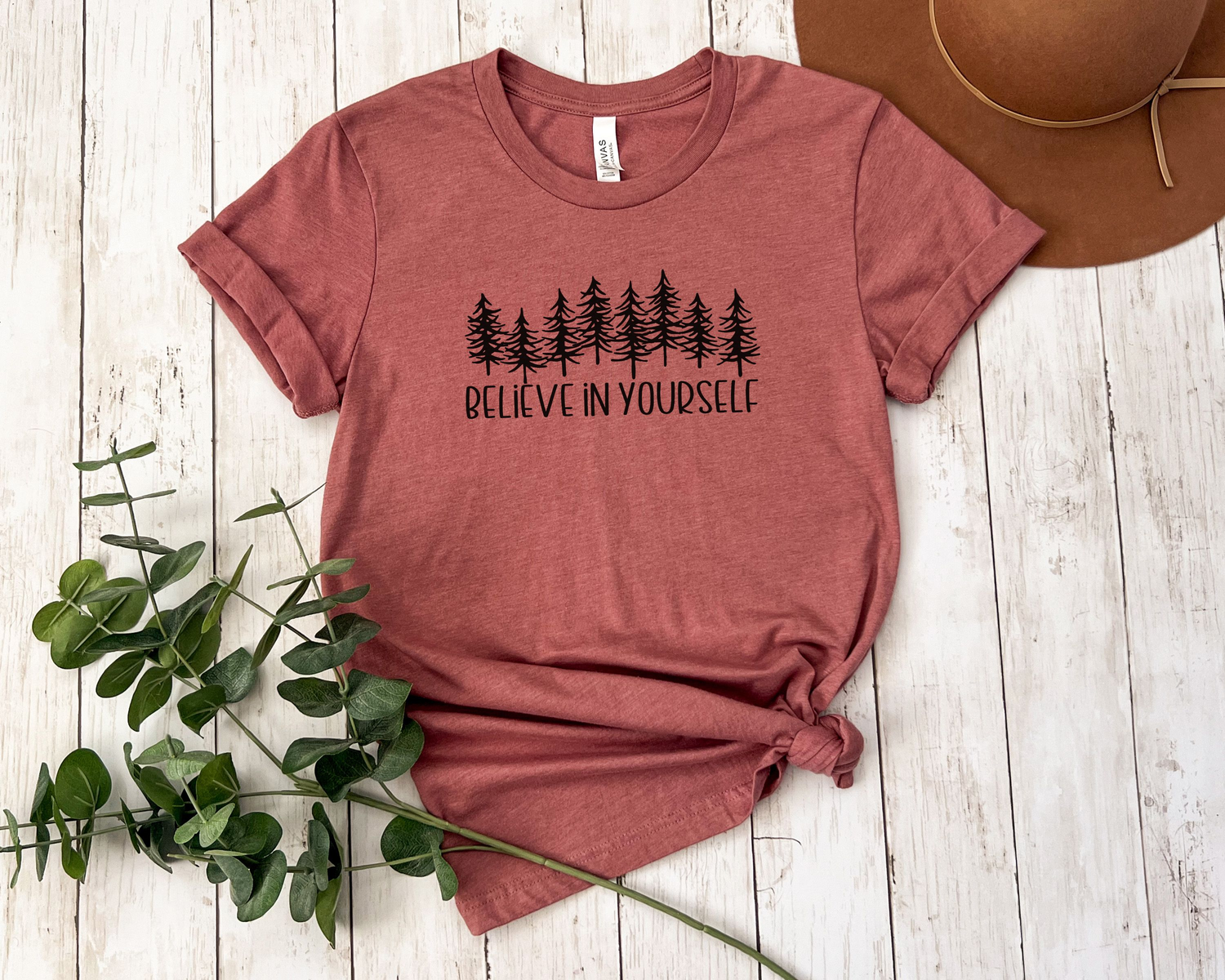 Mauve colored T-shirt laying flat with the words "Believe in Yourself" in all caps under a forest of trees graphic on the front.