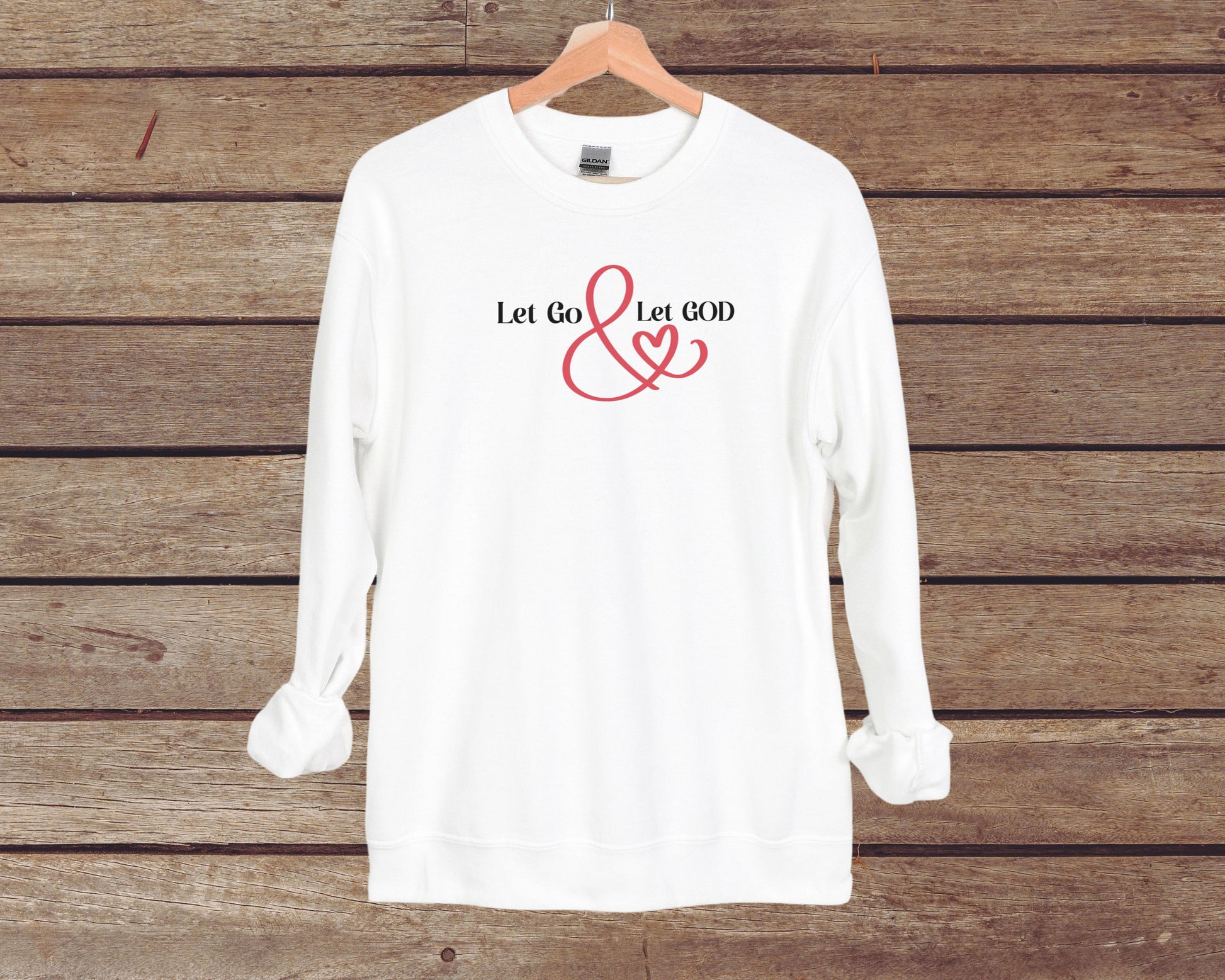 White crewneck sweatshirt with the phrase ‘Let Go & Let God’ in bold black text and a red ampersand forming a heart. Cozy, inspirational faith-based apparel for everyday wear.